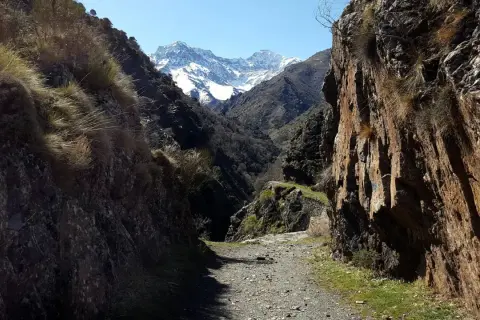 Camino de grava entre rocas altas, con pastos secos y montañas nevadas al fondo. Un sendero que parece la vereda de la estrella.