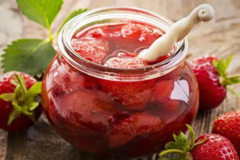 Ein Glas mit ganzen Erdbeeren und Sirup, bereit zum Erdbeeren einwecken. Frische Erdbeeren und Blätter liegen daneben.