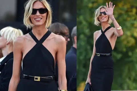 Elegancka kobieta w czarnej sukience i okularach przeciwsłonecznych, z subtelnymi złotymi akcentami, jak bransoleta i klamra paska.