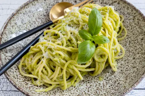 Cremige Avocado-Pasta mit frischem Basilikum und Parmesan auf einem rustikalen Teller, serviert mit schwarzem Besteck.