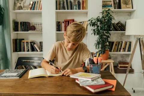 Un niño concentrado escribe en su cuaderno, rodeado de libros y útiles escolares, practicando dictados cortos para mejorar su ortografía.