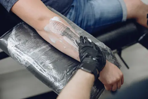 Ręka w czarnej rękawiczce nakłada piankę na świeży tatuaż. Całość jest owinięta folią spożywczą na tatuaż, chroniąc przed zakażeniem.