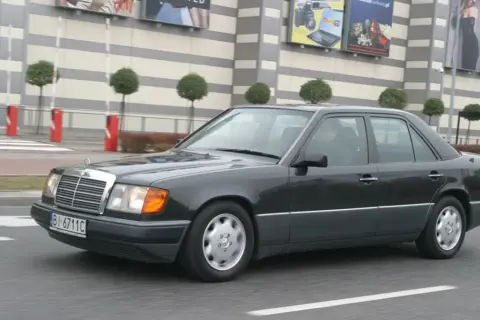 Ciemnoszary Mercedes W124 jedzie ulicą przed budynkiem z banerami.
