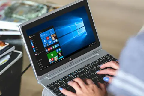 Kobieta pisze na laptopie z systemem Windows 10. W tle widać stosy czasopism, co sugeruje, że zadania w tle ochrony systemu są wykonywane podczas relaksu.