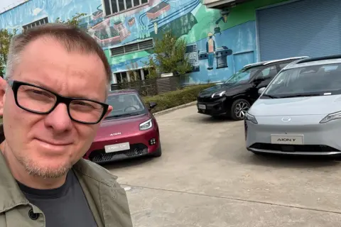 Mężczyzna w okularach pozuje do selfie na tle trzech samochodów marki AION.