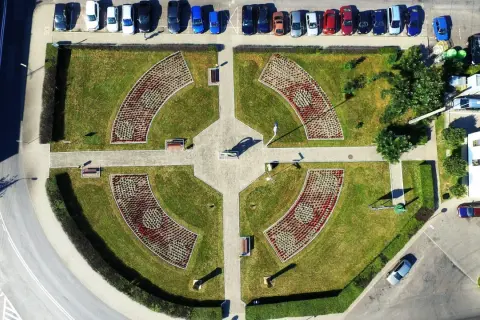 Piątek miasto środek Polski. Park miejski z klombami w kształcie łuków, alejkami i ławkami. W tle parking z samochodami.