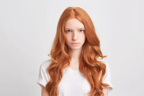 Dziewczyna z burzą rudych włosów, lekko falowanych, patrzy prosto przed siebie.