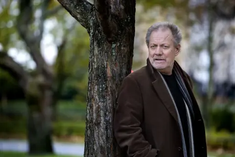 Harald Bovallius lutar sig mot ett träd i en park, klädd i brun rock och halsduk.