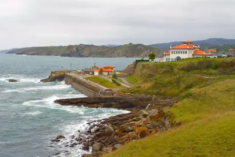 Costa de San Vicente de la Barquera a Comillas con edificios blancos y tejados naranjas, mar agitado y acantilados verdes.