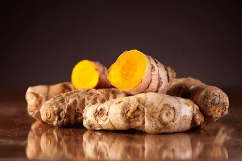 Ein Haufen frischer Kurkumawurzeln, eine davon aufgeschnitten, enthüllt ihr leuchtend orangefarbenes Inneres und die wertvollen Kurkuma Inhaltsstoffe.
