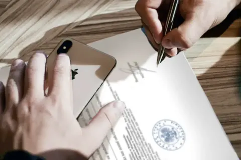 Dłoń trzyma iPhone'a na dokumencie, podczas gdy druga ręka podpisuje go, co może symbolizować poddanie się egzekucji.