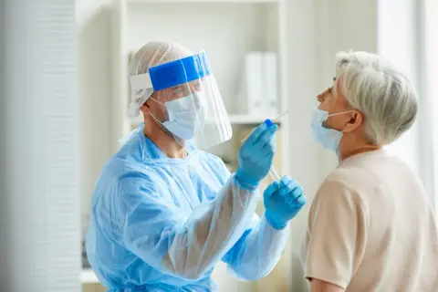 Arzt in Schutzkleidung nimmt bei älterer Frau einen Rachenabstrich. Testet man so auch auf MRSA, was ist das?