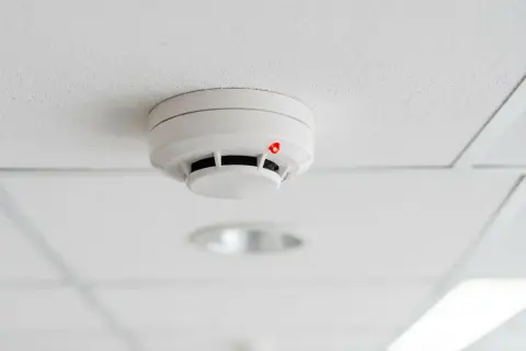 Détecteur de fumée blanc sur un plafond blanc, avec un voyant rouge allumé, suggérant un détecteur qui se déclenche tout seul.