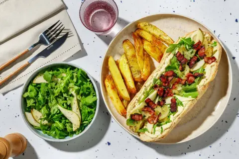 En härlig fransk mat-inspirerad måltid med en baguette fylld med krämig sås, ruccola, päron och knaprigt bacon, serverad med klyftpotatis och en fräsch sallad.