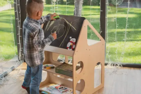 A young boy draws on a chalkboard easel, part of a Montessori backyard playhouse. He's erasing a drawing with a green eraser.