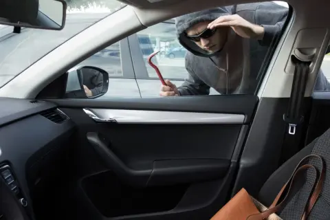 Un homme en capuche et lunettes de soleil tente un vol à la roulotte, outil rouge à la main, regardant à l'intérieur d'une voiture.