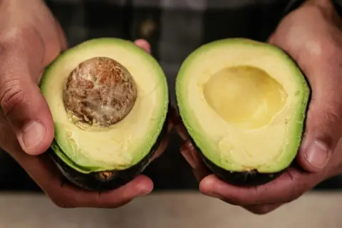 Dłonie trzymają połówki awokado. Jedna z nich ma pestkę, druga jest pusta. Idealne do guacamole, gdy wiesz, jak zmiękczyć awokado.