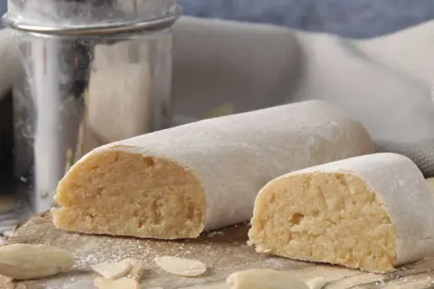 Pâte d'amande, c'est quoi ? Deux rouleaux de massepain, saupoudrés de sucre glace, sur une planche en bois avec des amandes effilées.