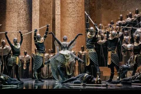 Scena z opery Aida: tancerze w czarnych strojach ze złotymi zdobieniami i maskami na twarzach, niektórzy z włóczniami, inni leżą na scenie.