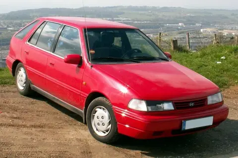 Czerwony Hyundai Pony z lat 90. stoi na brudnej drodze, z widokiem na dolinę i miasto w tle.