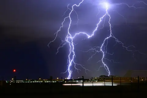 Un spectaculaire **photo orage** illumine le ciel nocturne au-dessus d'un aéroport. Des éclairs zèbrent l'obscurité, illuminant les bâtiments et les lumières de piste.
