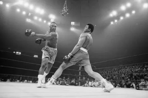 Two boxers clash in a dramatic moment. One out boxer throws a punch while the other dodges, the crowd watching intently.