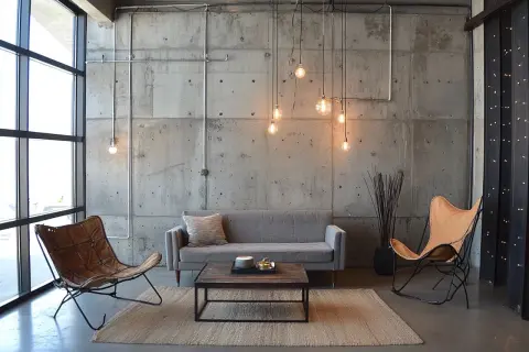 Salon w industrialnym stylu: szara kanapa, dwa fotele (jeden skórzany, drugi z brązowej skóry), drewniany stolik kawowy, betonowa ściana z widocznymi instalacjami i lampami.