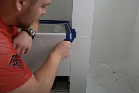 Mężczyzna w czerwonej koszulce przygotowuje się do tego, jak silikonować narożniki, oklejając je taśmą.