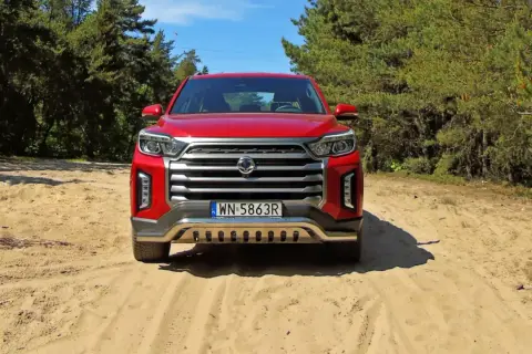 Czerwony Daewoo Musso na piaszczystej drodze wśród drzew.