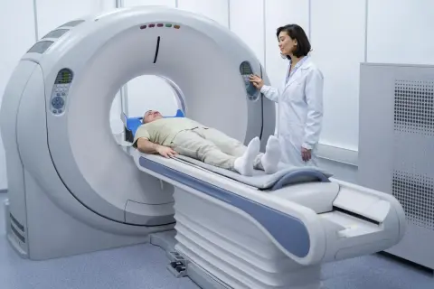 Pacjent leży na stole rezonansu magnetycznego, przygotowywany do badania. Lekarka w białym fartuchu obsługuje konsolę.