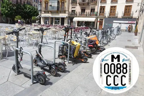 Patinetes eléctricos y bicicletas aparcados en una plaza. Se ve una pegatina de la DGT con la matricula CD.
