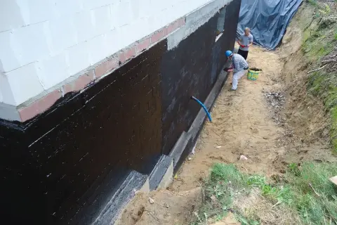 Dwóch mężczyzn wykonuje hydroizolację fundamentów czarną masą KMB.