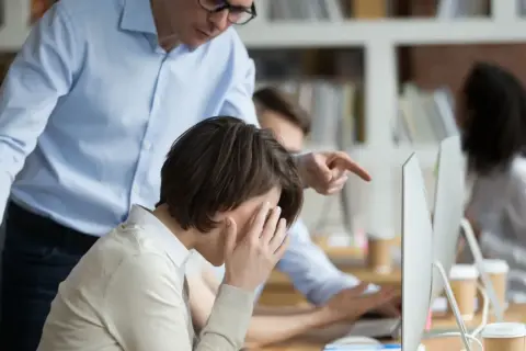 Przełożony wskazuje palcem na pracownicę, która zakrywa twarz dłońmi. Widać niewłaściwe zachowanie przełożonego wobec pracownika.