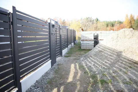 Schemat fundamentu pod bramę przesuwną. Widać nowoczesne ogrodzenie, materiały budowlane i hałdę żwiru.