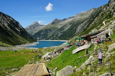 Ein idyllisches Bergpanorama mit einem türkisfarbenen See, umgeben von steilen Hängen. Hütten und Wanderer beleben das Bild, das an ein kleines Tibet im Zillertal erinnert.