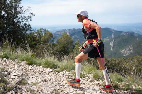 Uomo con bastoncini da trail running cos'è, corre su sentiero roccioso con vista panoramica su montagne e cielo azzurro.