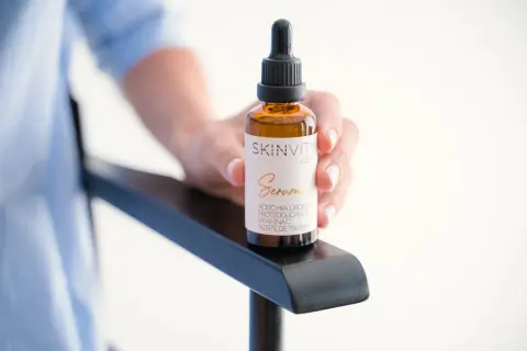 Mano sostiene un frasco de vidrio ámbar con gotero, etiquetado como "SKINVITY care Serum". Este booster es un sérum facial.