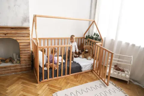 Two children play in a wooden playpen floor bed. One reads a book, the other stands, with a teddy bear nearby.