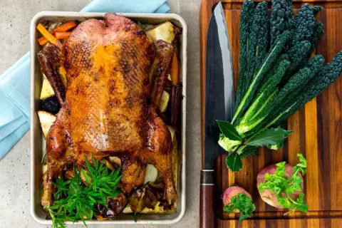 Helstekt anka med rotfrukter i ugnsform. Bredvid ligger en stor kniv, grönkål och rödbetor. Perfekt anka temperatur för en festmåltid.