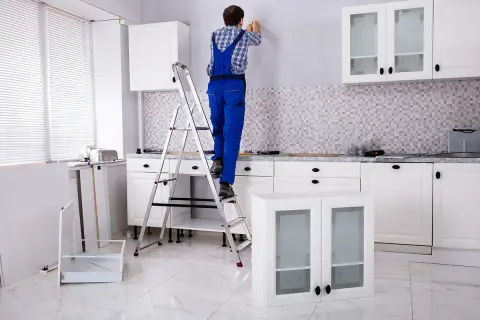 Un homme en salopette bleue est sur un escabeau, prêt à fixer un meuble au mur. La cuisine est en cours d'installation.
