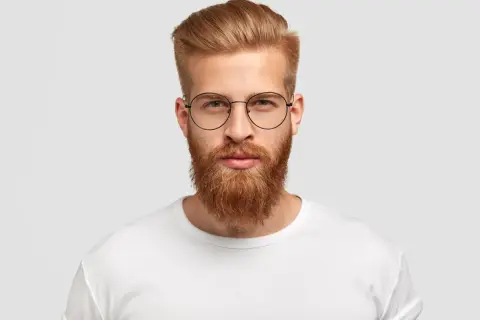 Un homme au visage rectangulaire, barbe rousse et lunettes rondes, porte un t-shirt blanc.