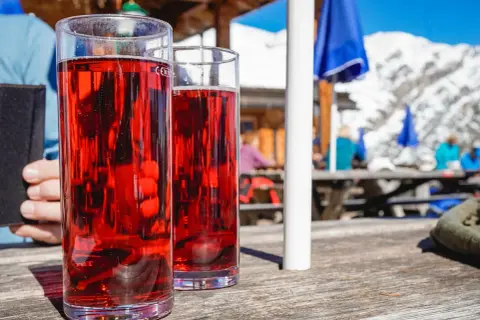 Zwei Gläser mit rotem Getränk, vielleicht ein Skiwasser-Rezept, stehen auf einem Holztisch. Im Hintergrund eine Skihütte mit Gästen.