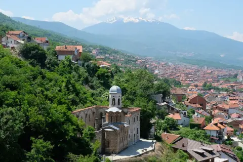 Eine malerische **Kosovo Landschaft** mit einer alten Kirche im Vordergrund, umgeben von üppigem Grün. Im Hintergrund erstreckt sich eine Stadt mit roten Dächern bis zu schneebedeckten Bergen.