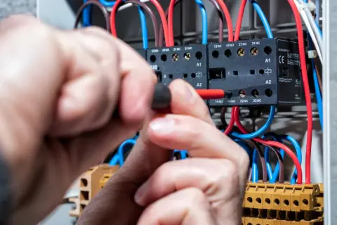 Ręce majsterkowicza przykręcają śrubę w skrzynce elektrycznej, gdzie widać przewody i styczniki. Kontrola napięcia prądu.