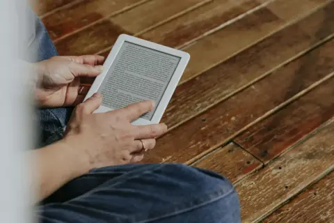 Une personne lit sur une tablette encre électronique, assise sur un plancher en bois.