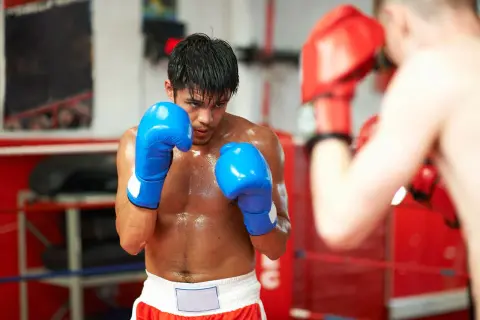 A boxer in blue gloves sweats during a tough boxing cardio workout in the ring, facing an opponent in red gloves.
