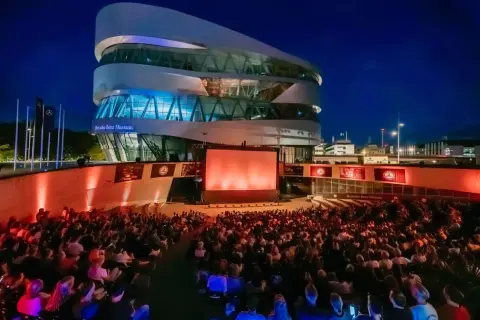 Ein großes Publikum genießt einen Filmabend beim Mercedes-Benz Museum Open Air. Die moderne Architektur des Museums bildet die beeindruckende Kulisse.