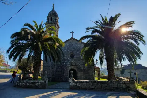 Iglesia de piedra con palmeras y sol brillante. Un lugar tranquilo que ver en Rianxo, ideal para pasear.