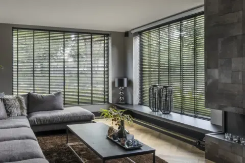 Nowoczesny salon z szarym narożnikiem i stolikiem kawowym. Duże okna z czarnymi żaluzjami wewnętrznymi, które dodają elegancji.