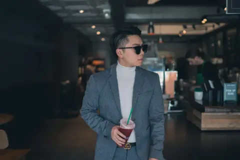 Mann im grauen Anzug mit weißem Rollkragenpullover und Sonnenbrille, hält Getränk. Das ist smart casual.